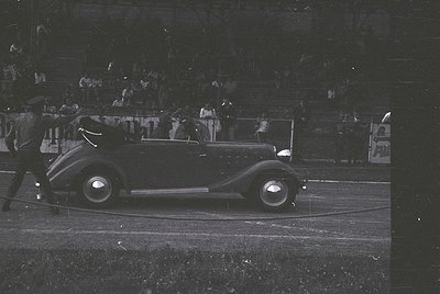 Classic 1930s convertible race car on a track, surrounded by spectators. Streamlined bodywork and dual headlights highlight v...