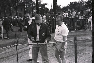 Mid-20th century outdoor event with rope barrier separating crowd from participants. Two men in casual workwear (one in a hat...