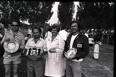 Four individuals pose outdoors holding trophies, likely from a 1970s sports or cultural event. The man on the left wears sung...