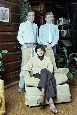 Family portrait in a rustic, wood-paneled interior, likely mid-20th century (1960s–1970s). Three individuals pose: two standi...