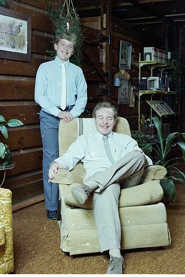 Vintage indoor portrait featuring a seated man in a light-colored suit and tie, relaxed in a vintage armchair, with a standin...