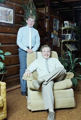 Two men pose indoors in a rustic wooden-paneled room, likely mid-20th century. The seated man wears a light-colored suit with...