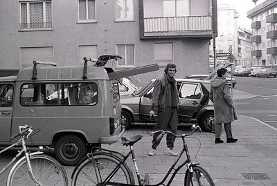 Mid-20th century urban street scene with vintage Volkswagen van carrying surfboard. Two men in casual 1970s attire—one standi...