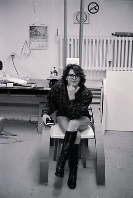 A woman in 1970s-style glasses and patterned coat sits on a white stool in an industrial office, holding a phone receiver. Su...