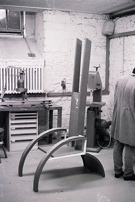 Industrial workshop featuring mid-century woodworking tools. A person operates a band saw with a curved wooden piece on the t...