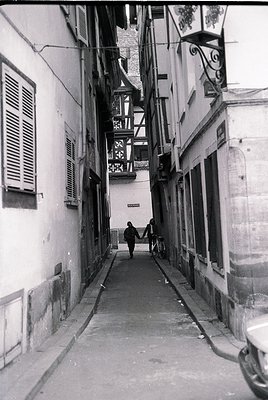 Narrow alleyway flanked by mid-20th-century European timber-framed buildings, featuring shuttered windows and decorative iron...