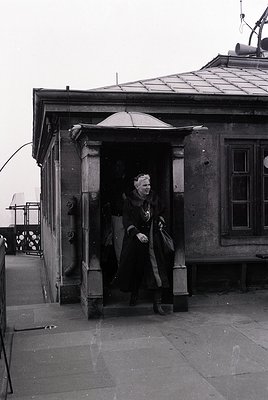 Mid-20th century black-and-white photo of a woman exiting a stone building’s arched entrance, holding a large umbrella and co...