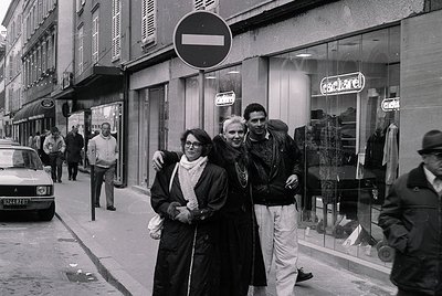 Mid-20th century street scene in Paris, featuring three central figures in 1960s fashion: a woman in a dark coat and scarf, a...