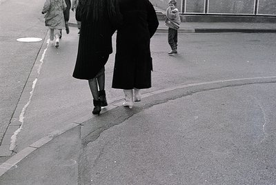 Black-and-white street scene featuring two women in long, floor-length coats and ankle boots crossing a curved sidewalk. One ...