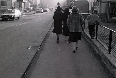 Four individuals in 1970s-era attire walk away from the camera on a paved sidewalk beside a road. The woman in the center wea...
