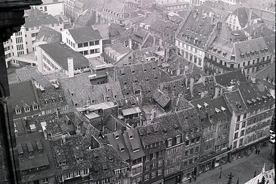 Dense urban rooftops and chimneys in a European city, likely post-WWII, showing bomb damage and reconstruction. Sloped slate ...