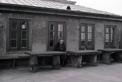 Black-and-white photo of elderly woman seated on concrete bench outside weathered concrete building with large rectangular wi...