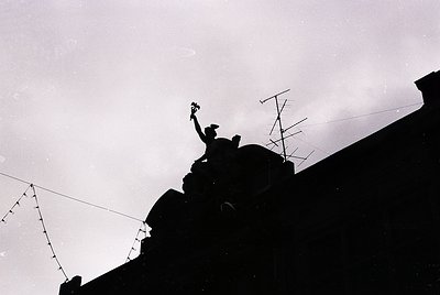 Silhouetted figure atop a rooftop, raising a small object (likely a flag or flower) against a cloudy sky. Urban setting with ...