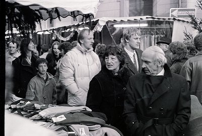 Crowded indoor market scene with festive garlands, featuring mid-20th century clothing (1960s-70s). Elderly man in overcoat a...