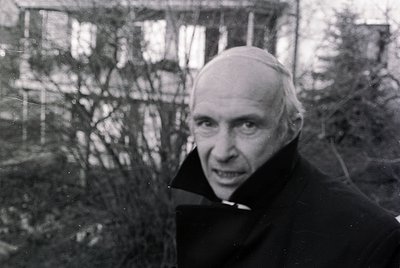Black-and-white portrait of an elderly man in a clerical collar, likely a priest, outdoors with blurred winter trees in backg...