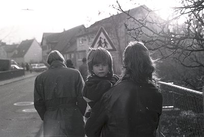 Black-and-white street scene featuring three adults walking with a child in a rural or small-town setting. The adult holding ...