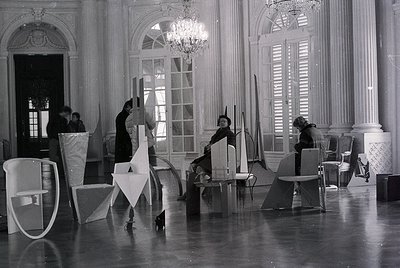 Mid-century modern furniture design studio in a grand, classical interior. Four individuals collaborate with geometric, minim...