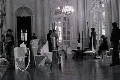 Black-and-white shot of a grand interior featuring geometric set pieces and crew positioning in a spacious, ornate room. Orna...