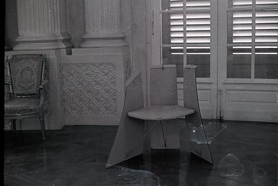 Vintage black-and-white interior of a grand, abandoned room with ornate plasterwork and shuttered windows. Broken glass and a...