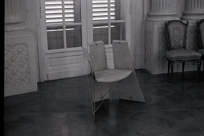 Vintage folding chair placed beside an ornate, upholstered armchair in a grand, dimly lit room. Decorative plasterwork and sh...