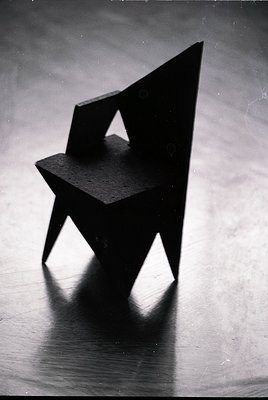 Minimalist geometric chair with sharp, angular design—likely mid-century modern. Dark material (wood or plastic) with clean, ...