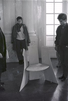 Vintage black-and-white photo of two individuals examining a modernist, angular stool in a grand interior. Ornate plasterwork...