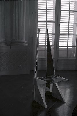 Minimalist geometric chair with angular, reflective metal frame positioned in a dimly lit interior. Ornate plaster columns an...