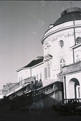 Neoclassical building corner with ornate dome and balustrade, likely 19th-century European architecture. Group of people asce...