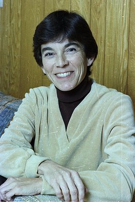 Portrait of a smiling individual in a vintage-style sweater, seated indoors with wooden paneling. Lighting suggests a mid-20t...