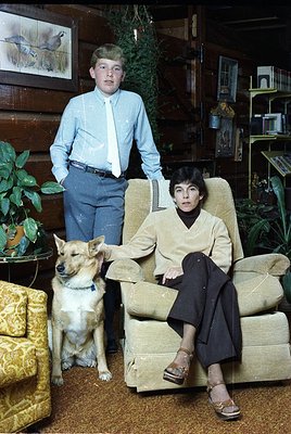 Vintage indoor portrait featuring a woman in a beige sweater and black trousers seated on a vintage armchair, a young boy in ...