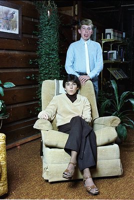 Mid-century interior featuring a woman in a beige blazer and black trousers seated in a vintage armchair, flanked by a standi...