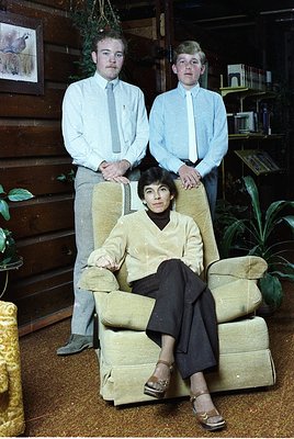 Vintage family portrait in a wood-paneled interior, likely 1960s–1970s. Three individuals pose: two standing men in light-col...