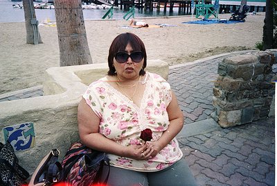 A woman in a floral blouse and sunglasses sits on a stone bench beside a seaside promenade, holding a red rose. Beachgoers re...