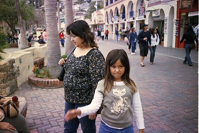 A woman and child stroll through a Mediterranean-style plaza with arched storefronts and palm trees. The woman wears a black ...