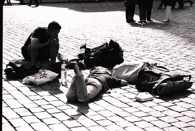 Two individuals rest on cobblestone pavement, surrounded by backpacks and personal items. The person on the left kneels, lean...