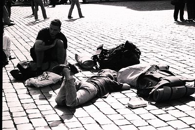 Two travelers rest on cobblestone pavement, surrounded by backpacks and luggage. One sits cross-legged, the other reclines. M...