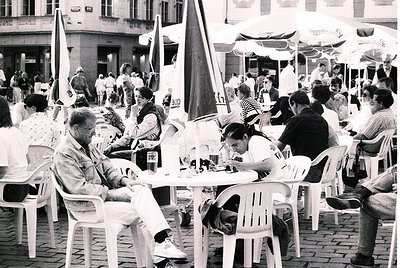 Vintage black-and-white street café scene with plastic chairs and tables under striped umbrellas. Mid-20th-century European u...