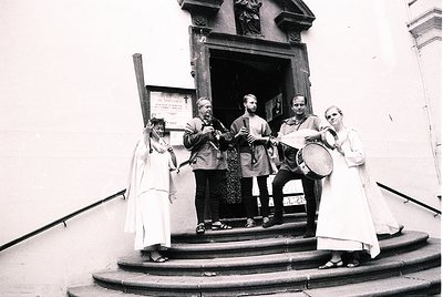 Medieval-themed street performance group in period costumes—flute, lute, drum, and lyre—posing outside a building entrance. C...