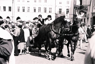 Horse-drawn carriage with two black horses in a bustling European street, mid-20th century. Crowd in vintage attire, multi-st...