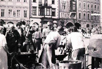 Black-and-white street scene featuring a lively outdoor market in a European city square, likely 1960s–1970s. Crowd gathers a...