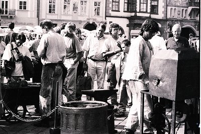 Crowded outdoor street market scene featuring blacksmithing demo—artisans work with an anvil and bellows, surrounded by onloo...