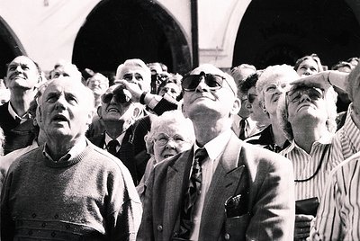 Crowd of elderly men in 1960s-70s attire, gathered indoors under arched stone ceilings, gazing upward with focused expression...