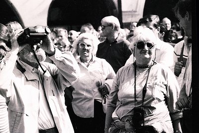 Vintage black-and-white photo of elderly individuals in a crowded, outdoor public space, likely a 1970s-1980s European city. ...