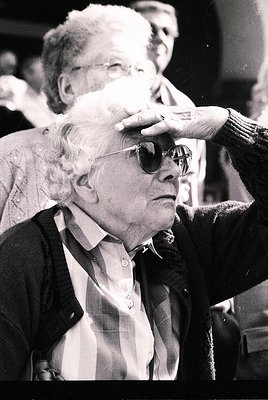 Tight close-up of an elderly woman in mid-20th century attire, wearing round sunglasses and a checkered jacket, with a hand a...