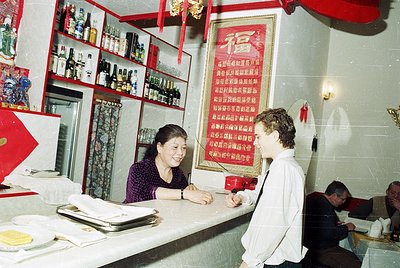 Vintage café interior with mid-century decor: woman in apron behind counter, man in white shirt ordering. Bottles, posters, a...