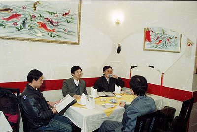 Four men in formal attire gather around a table in a dimly lit, traditional-style restaurant. Decorative koi fish paintings a...