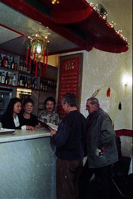 Vintage bar scene with five patrons at a counter adorned with red vinyl wallpaper and Chinese characters. Bottles line shelve...