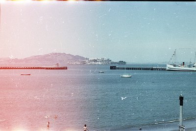 Vintage seaside pier extending into calm waters, flanked by small boats. Distant urban coastline with low-rise buildings and ...