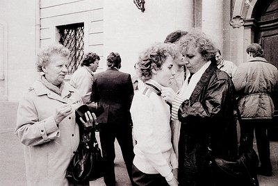 Black-and-white candid of three women in mid-conversation outside a building with classical architectural details. Women wear...