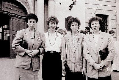 Four women in 1970s-era blazers and blouses pose outdoors near a grand, arched doorway. Formal attire suggests a professional...